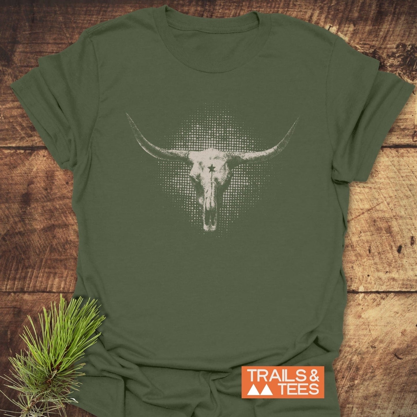 Buffalo Skull Texas T-Shirt on wooden surface, featuring a white steer skull graphic. Includes pine branch detail and TRAILS & TEES logo.