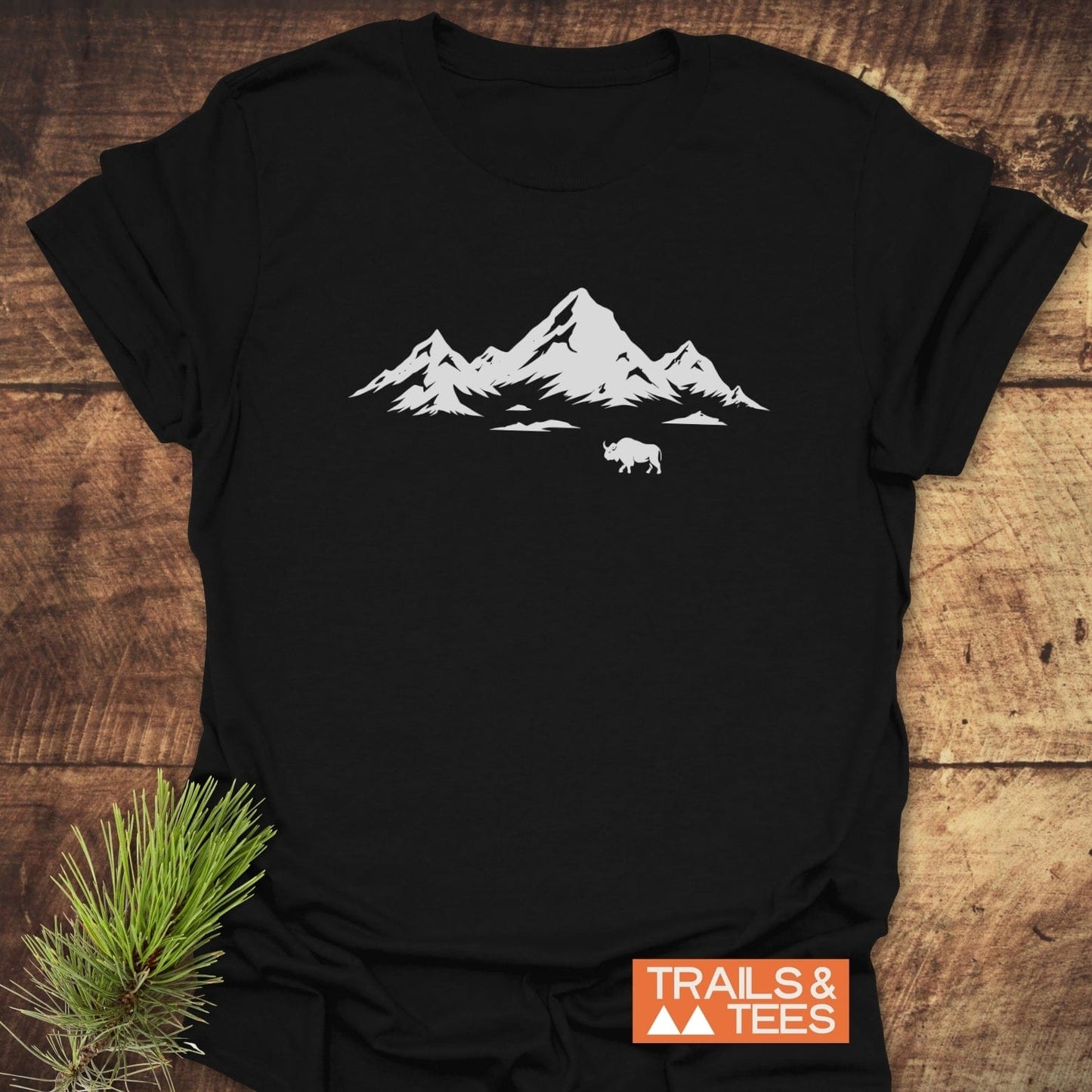 Mountains And Buffalo T-Shirt featuring a mountain silhouette and shaggy bovine print on soft cotton, laid on wood with pine sprig for outdoorsy touch.