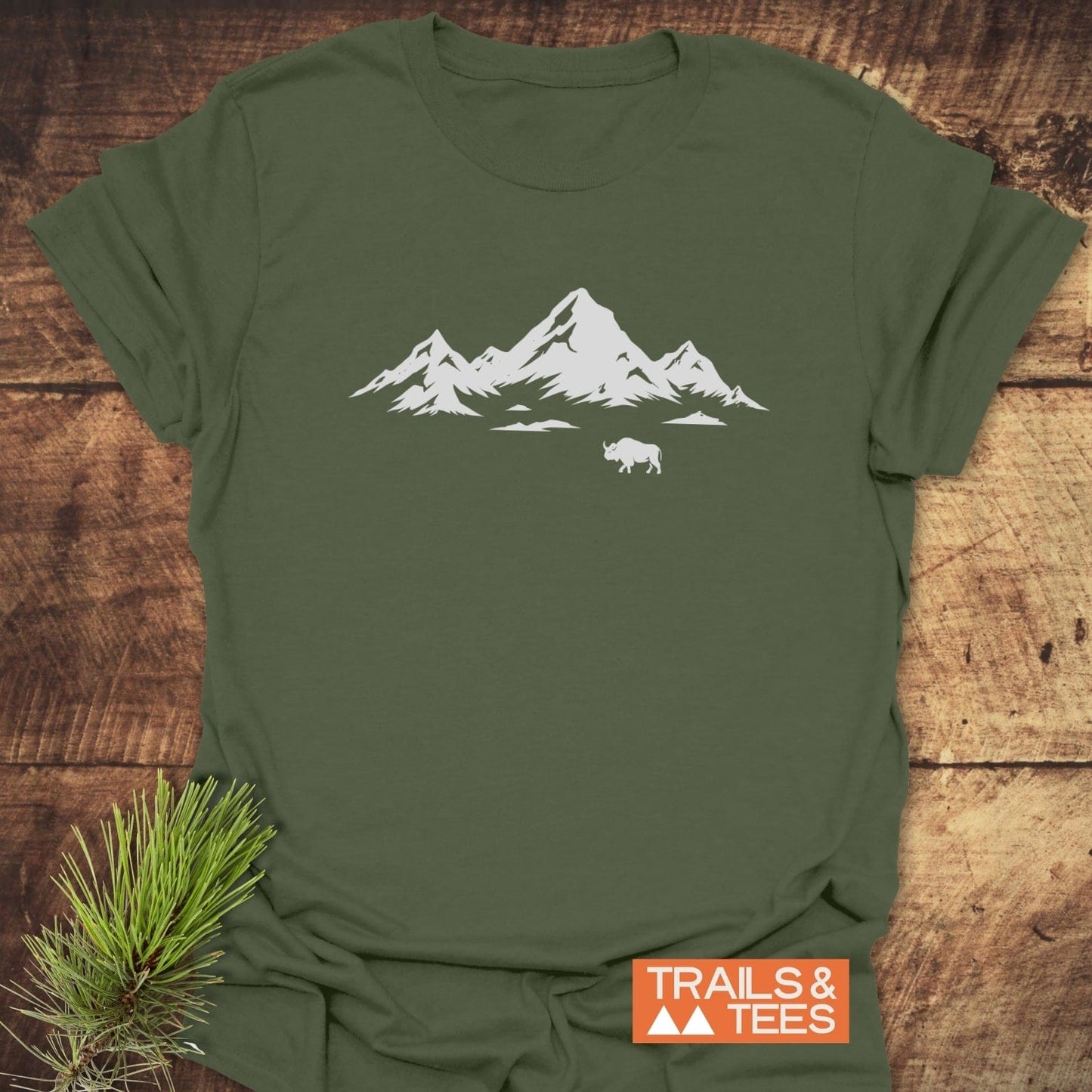 Mountains And Buffalo T-Shirt featuring a white mountain and bison graphic on an olive-green background, laid on wooden surface with a pine sprig.