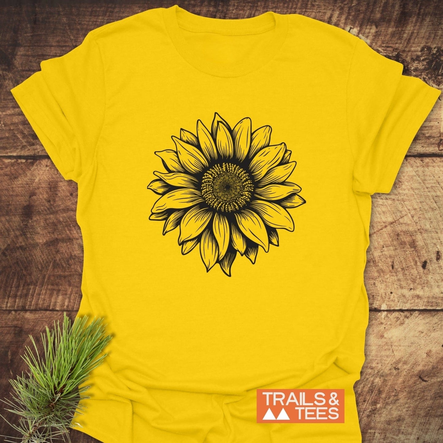 Sunflower T-Shirt on wood surface, featuring a black line-art sunflower. Includes a spruce branch for styling and TRAILS & TEES logo in the corner.