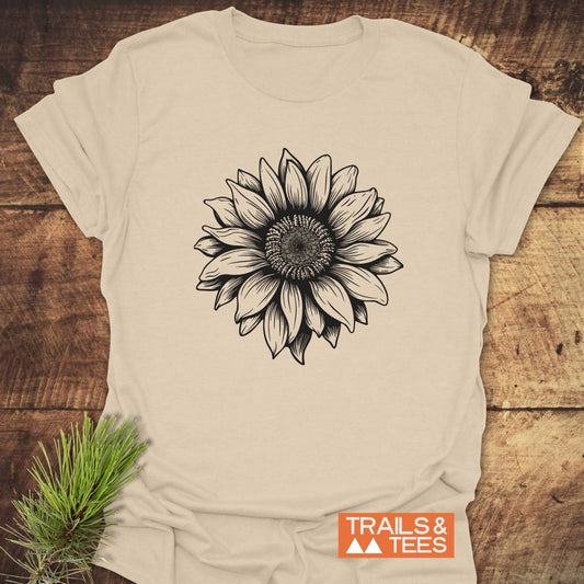 Light-beige Sunflower T-Shirt with a black sunflower drawing, crafted from soft cotton, laid flat. Features TRAILS & TEES logo and pine sprig beside it.