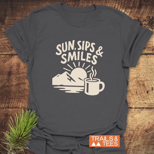 Sun, Sips, Smiles T-Shirt featuring a cream graphic of sunrise over mountains, with a steaming mug, on a charcoal-gray tee beside a pine bough.
