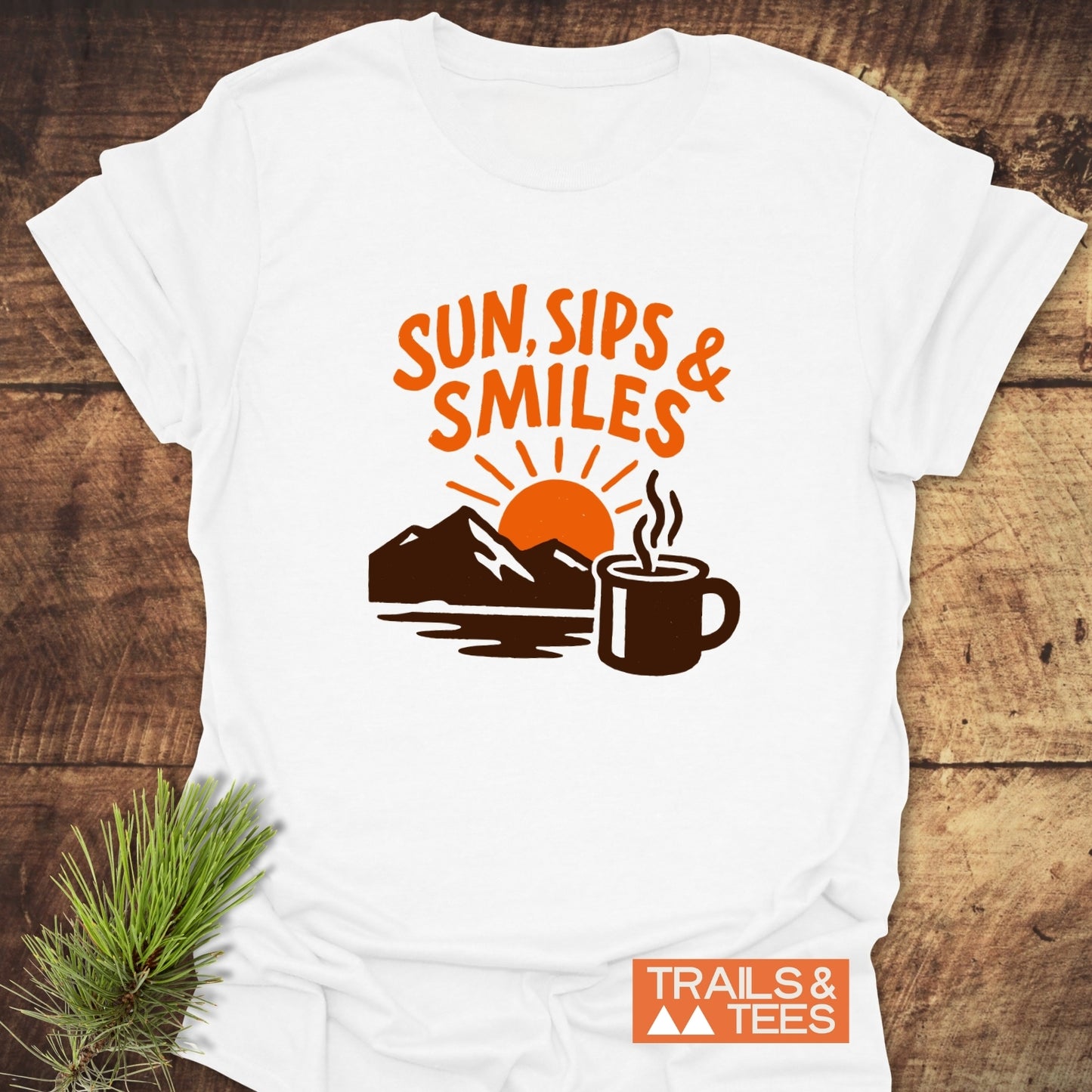 Sun, Sips, Smiles T-Shirt featuring a rising sun, mountains, and mug graphic. Crafted from soft cotton for comfort and adventure. Trails & Tees logo visible.