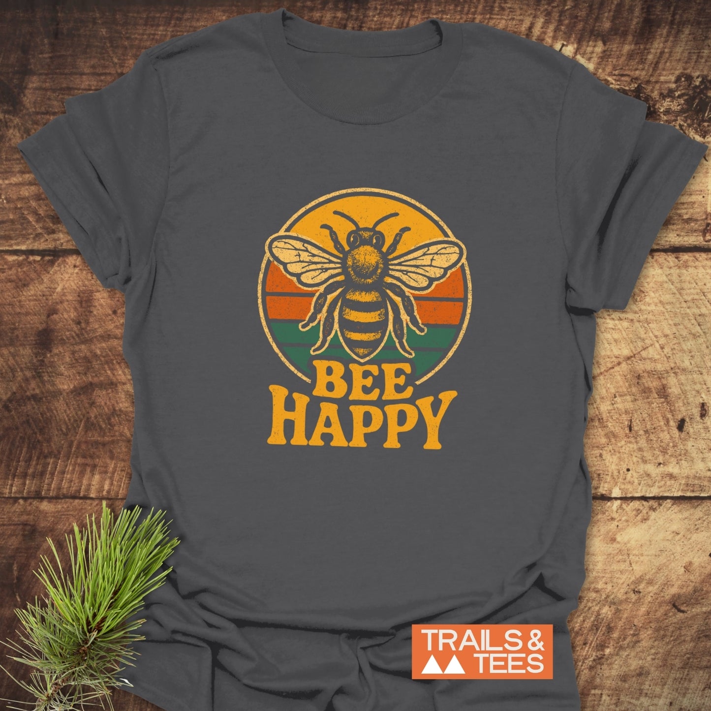 Bee Happy T-Shirt featuring a bee graphic and slogan, laid on wood with pine sprig. Soft, durable ring-spun cotton, perfect for outdoor adventures.