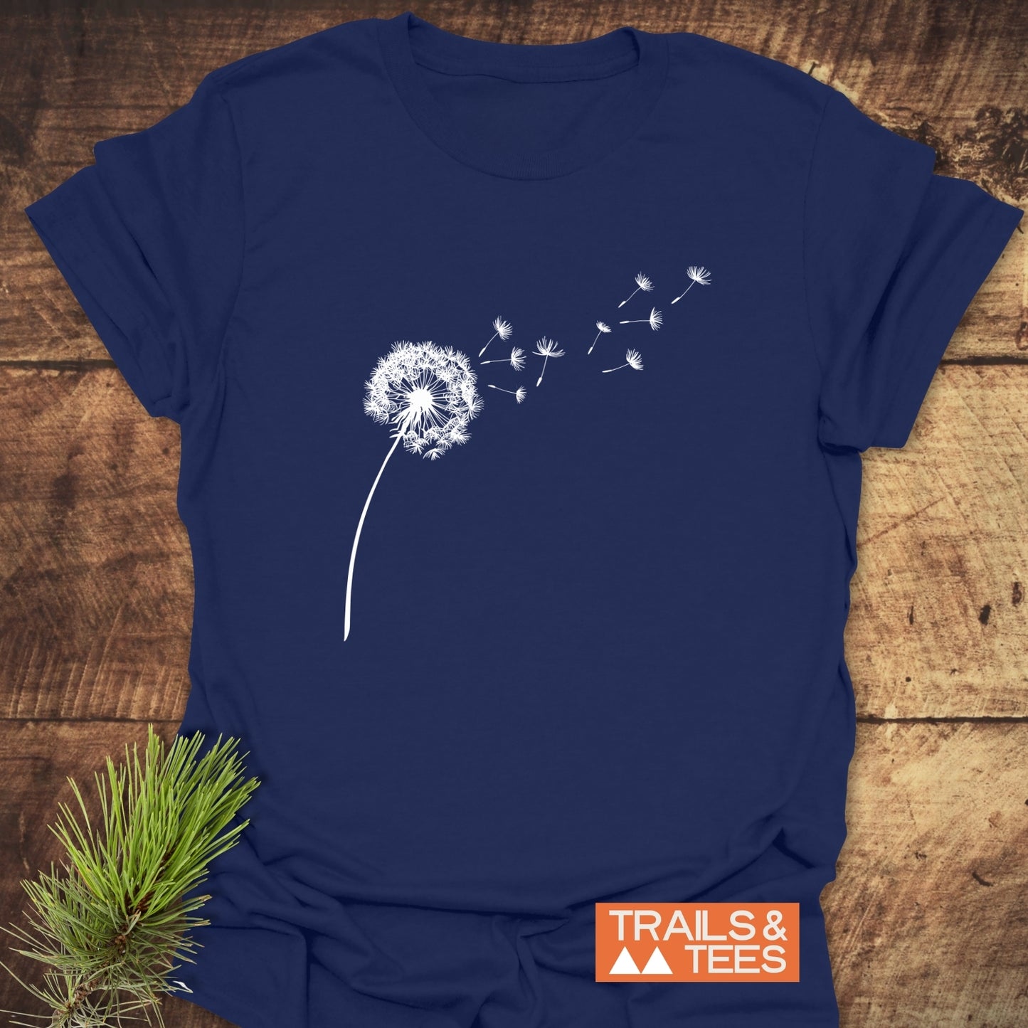 Navy-blue Dandelion T-Shirt with a white dandelion graphic, TRAILS & TEES logo, laid on wood, showcasing outdoor-inspired design and comfort.