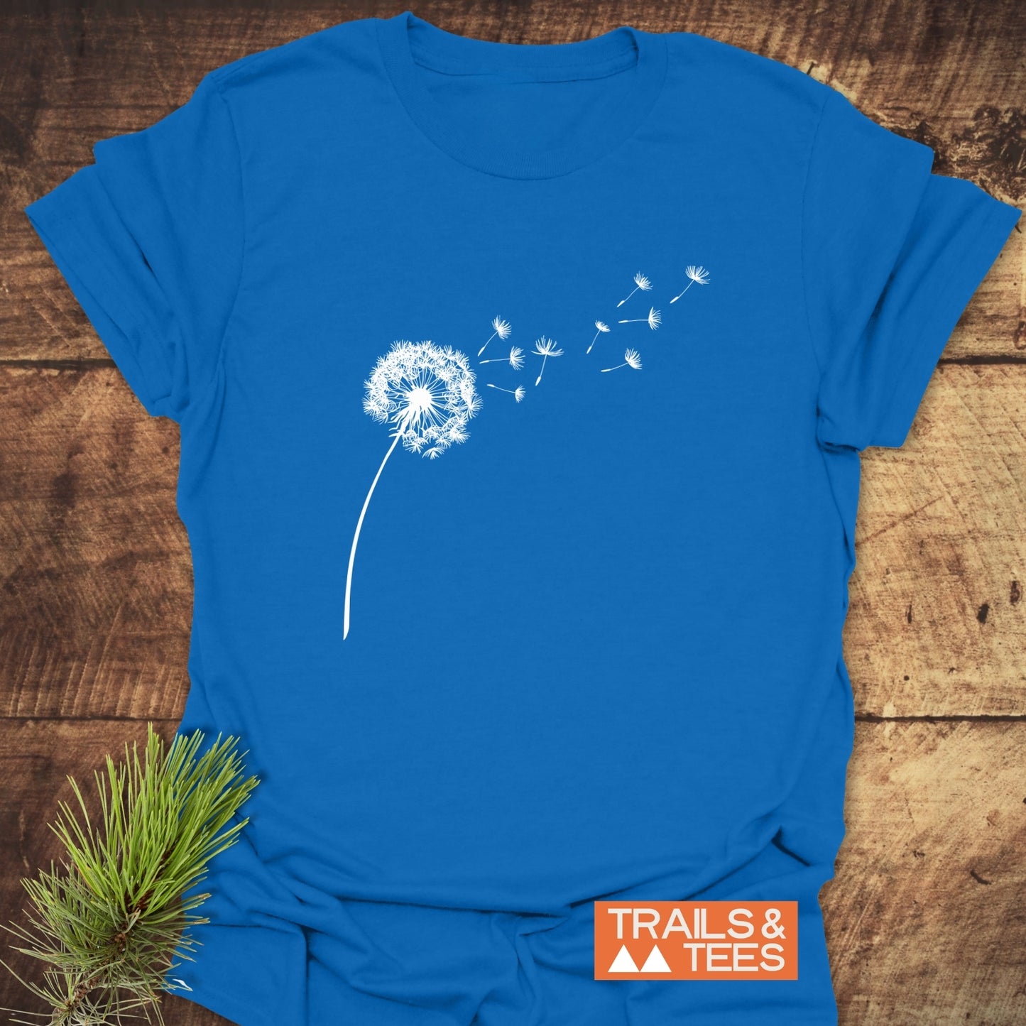 Dandelion T-Shirt featuring a white dandelion print on a blue fabric, showcasing a TRAILS & TEES logo and pine sprig detail for an outdoor-inspired look.