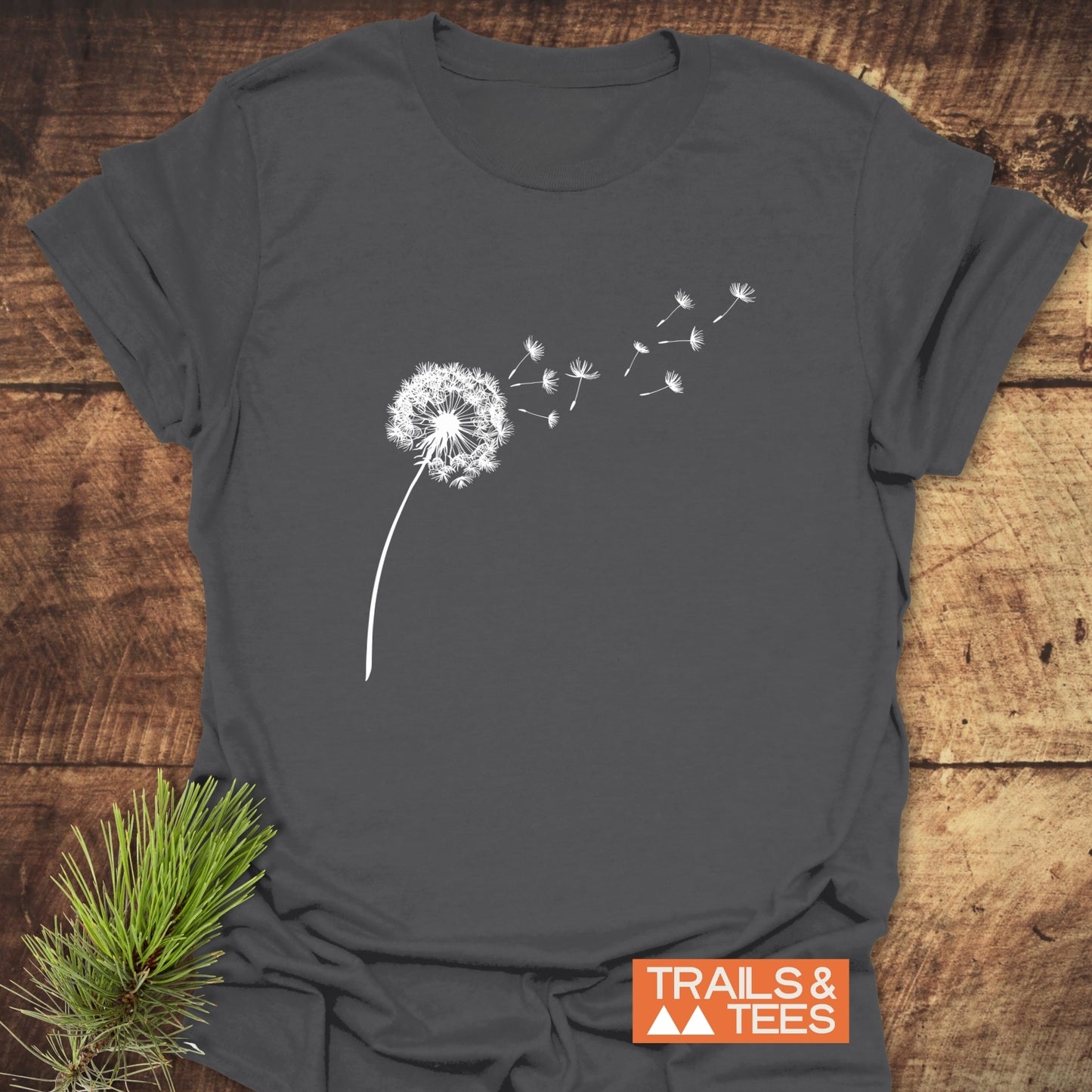 Dandelion T-Shirt featuring a white dandelion silhouette with seeds blowing, on a charcoal-gray fabric, highlighting the TRAILS & TEES logo, laid on a wooden surface.