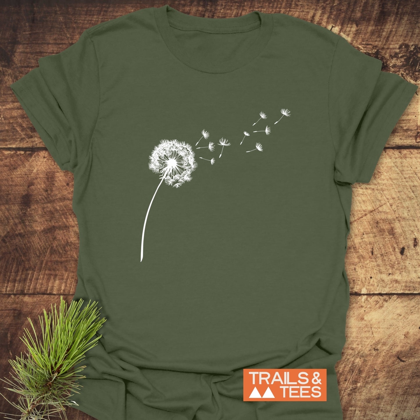 Dandelion T-Shirt featuring a white dandelion design, laid on wood with pine sprig, highlighting durable, trail-ready comfort from Trails & Tees.