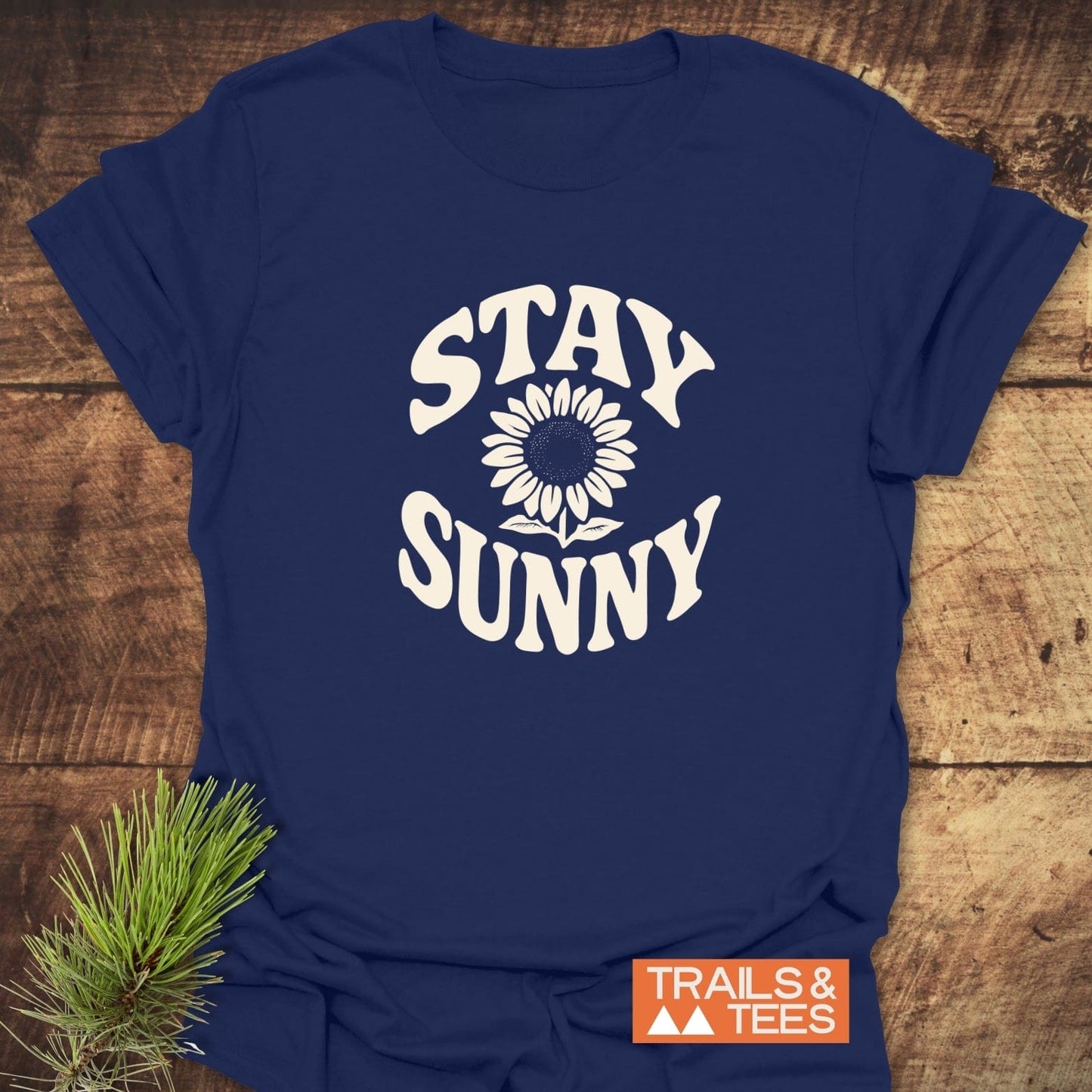 Stay Sunny T-Shirt with STAY SUNNY text and sunflower graphic, featuring a TRAILS & TEES logo; laid on wood with pine sprig.