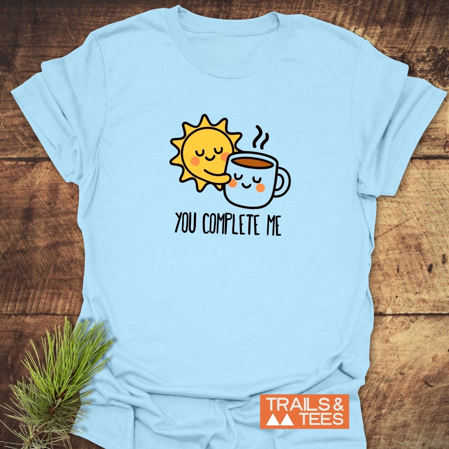 You Complete Me - Sun & Coffee T-Shirt with cartoon sun hugging coffee mug, 'TRAILS & TEES' logo, pine sprig, crafted for comfort and everyday adventure.