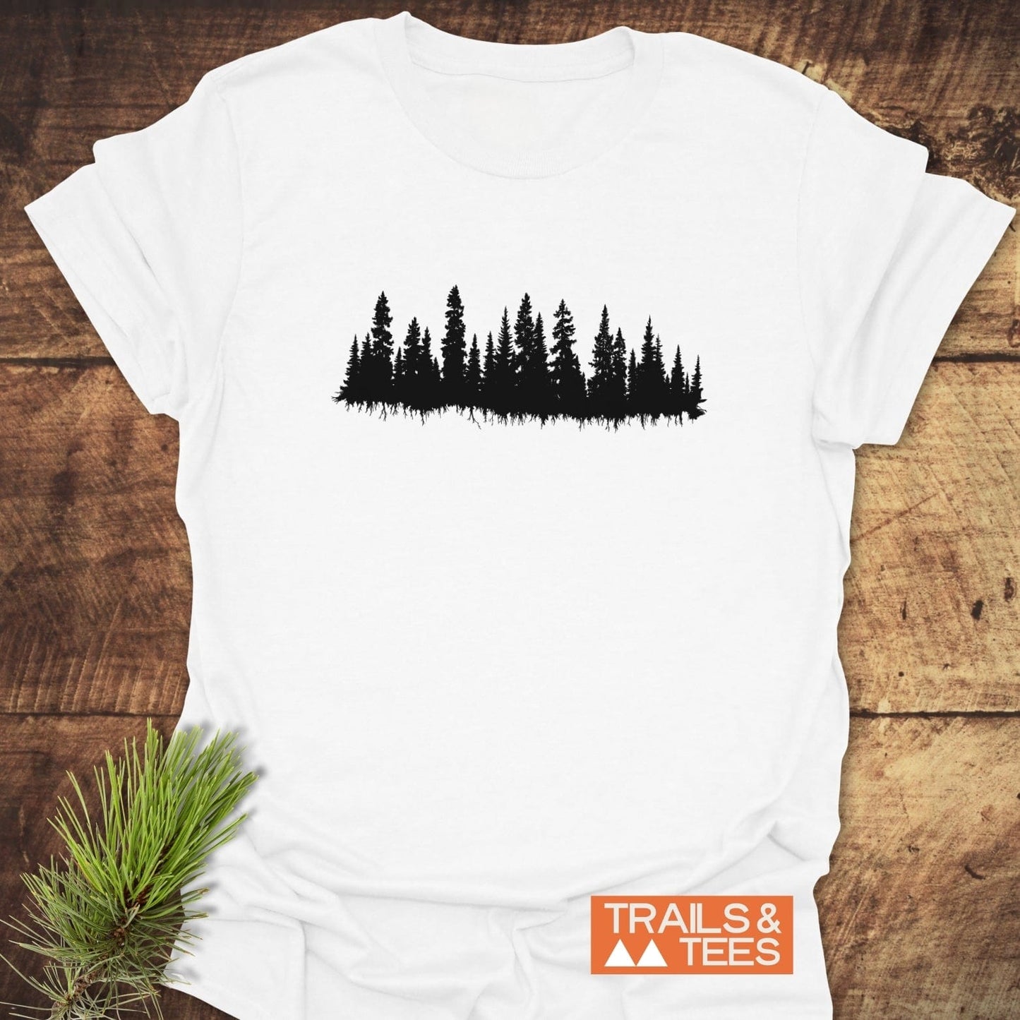 Trees Silhouette T-Shirt featuring a black pine forest design on white cotton, laid on a wooden surface with TRAILS & TEES logo and pine twig accent.