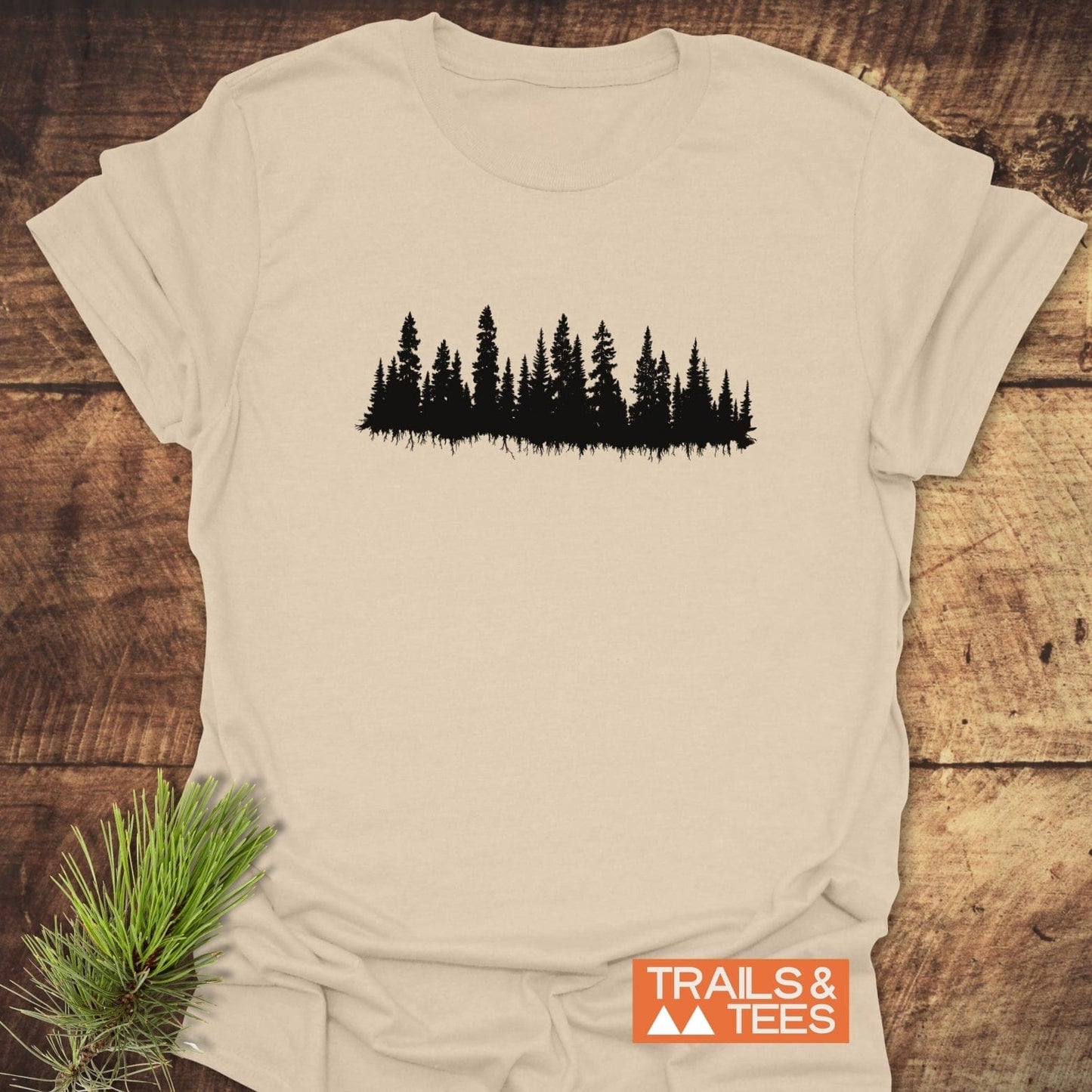 Cream-colored Trees Silhouette T-Shirt featuring a black pine-tree design, with an orange TRAILS & TEES logo, laid on a wooden surface.