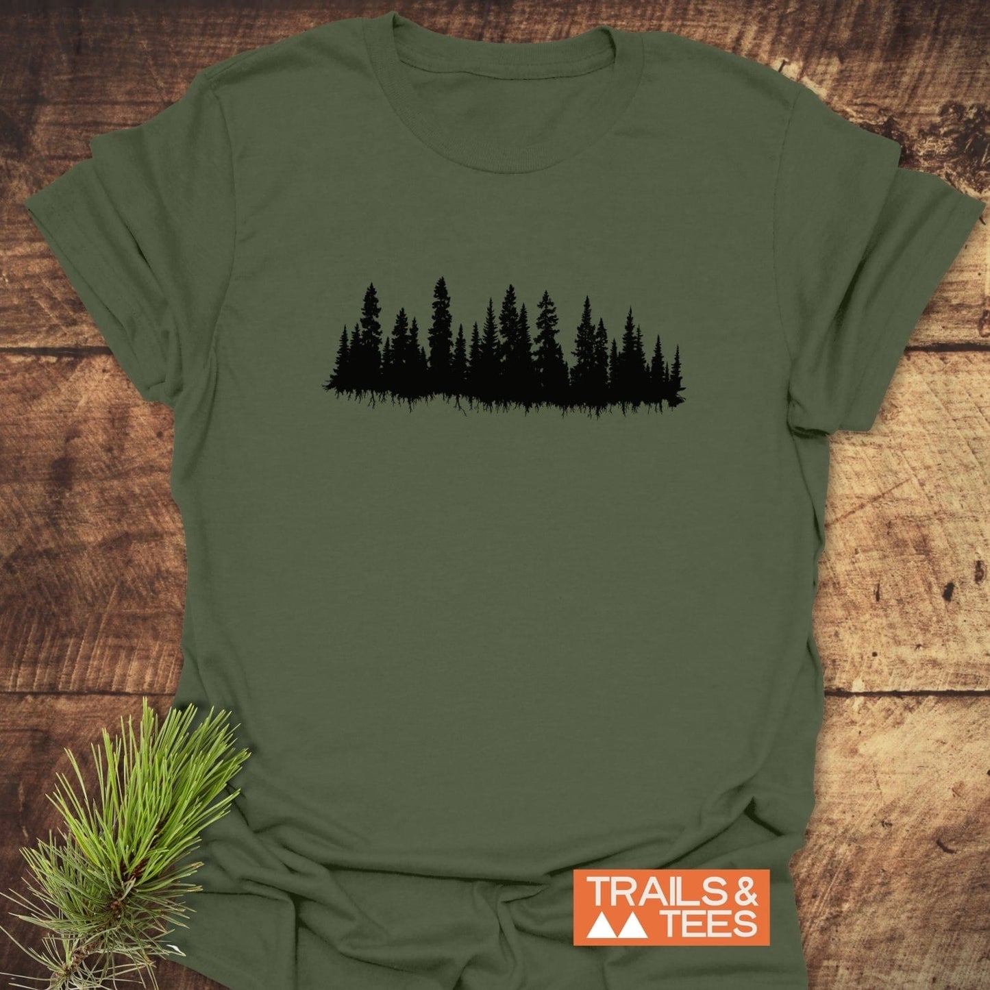 Trees Silhouette T-Shirt on wood surface, featuring a black forest treeline design. Includes evergreen sprig and TRAILS & TEES logo nearby. Crafted for outdoor enthusiasts.