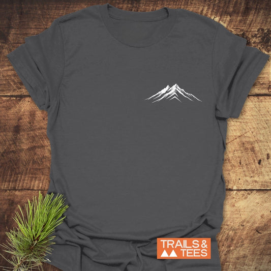 Mountains Logo T-Shirt featuring a minimalist mountain design on charcoal fabric, laid flat with pine sprig and TRAILS & TEES logo.