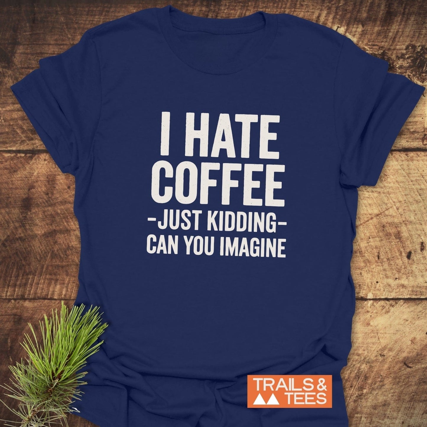 Navy blue I Hate Coffee T-shirt with white text, orange Trails & Tees logo. Laid on wood, pine sprig beside. Soft, durable cotton.