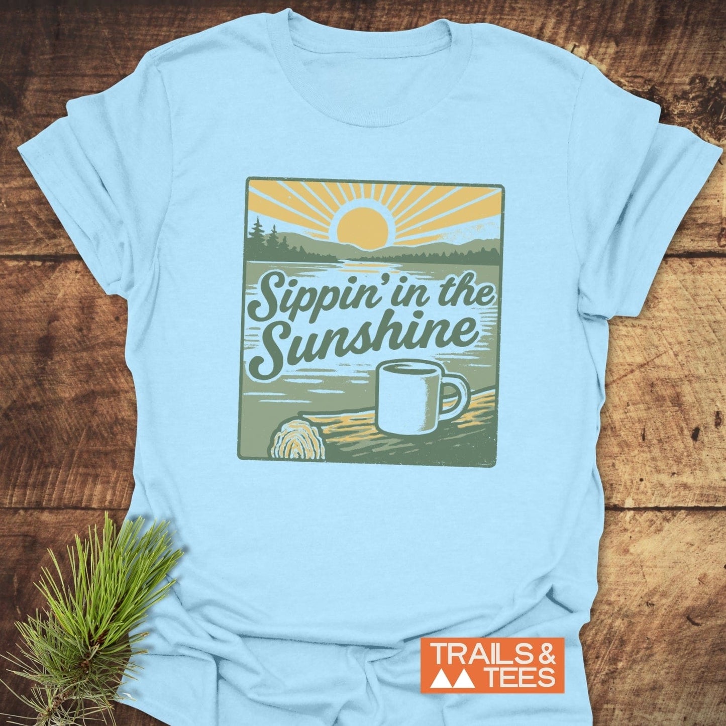 Sippin' In The Sunshine T-Shirt with sunrise graphic, coffee mug, and TRAILS & TEES label, offering comfort and adventure-ready design.