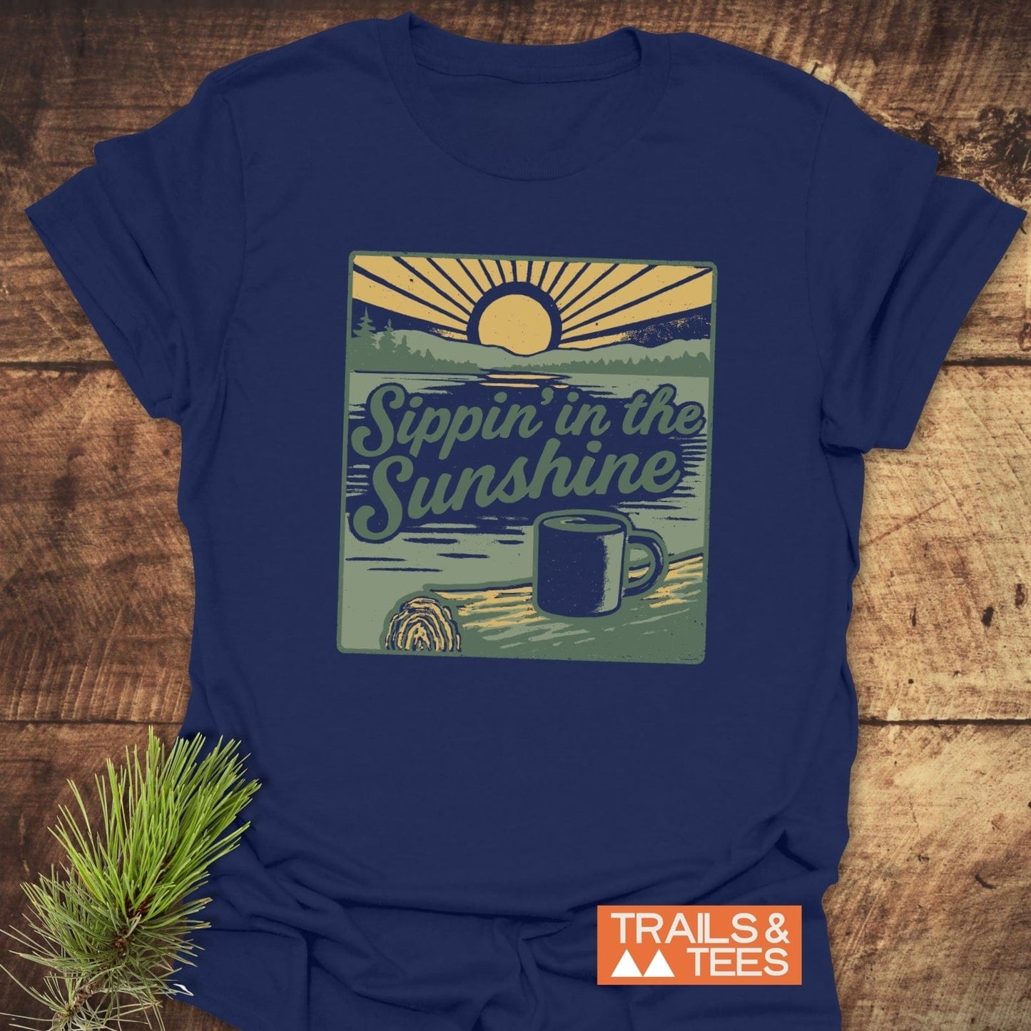Navy blue Sippin' In The Sunshine T-shirt with a lake sunrise graphic, set on wood, featuring a Trails & Tees logo and pine sprig.
