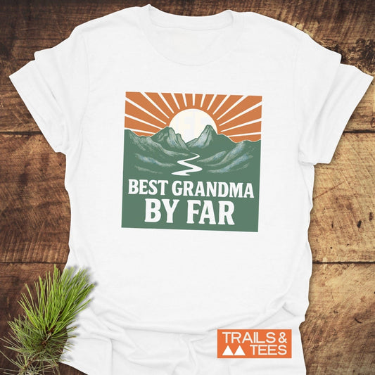 Best Grandma By Far T-Shirt featuring a stylized mountain graphic, crafted from soft cotton for comfort, laid on rustic wood with Trails & Tees logo.