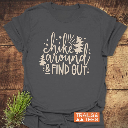 Hike Around And Find Out T-Shirt with off-white lettering and pine tree design, showcasing outdoor spirit on soft, comfortable ring-spun cotton, laid on wooden surface.