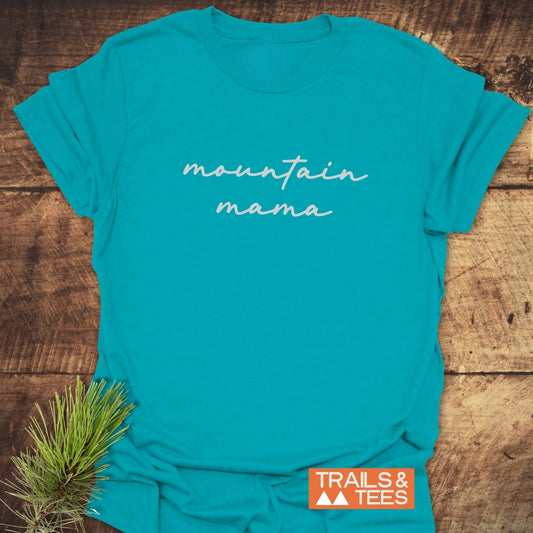 Mountain Mama T-Shirt on rustic wood, featuring mountain mama in white script. Includes orange TRAILS & TEES logo and styled with a sprig of pine.