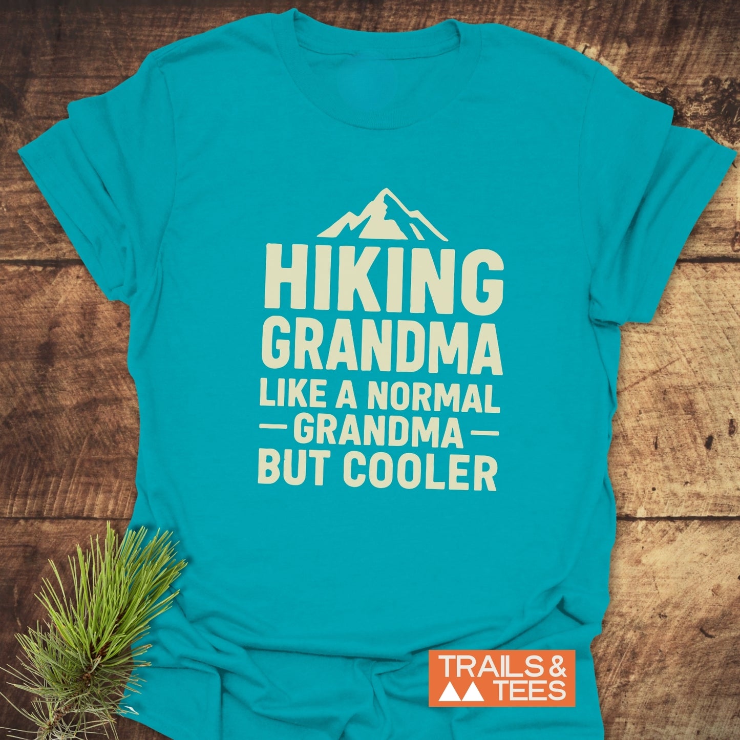 Teal Hiking Grandma T-Shirt with mountain graphic and slogan, featuring TRAILS & TEES logo. Laid on wood with pine needles nearby.