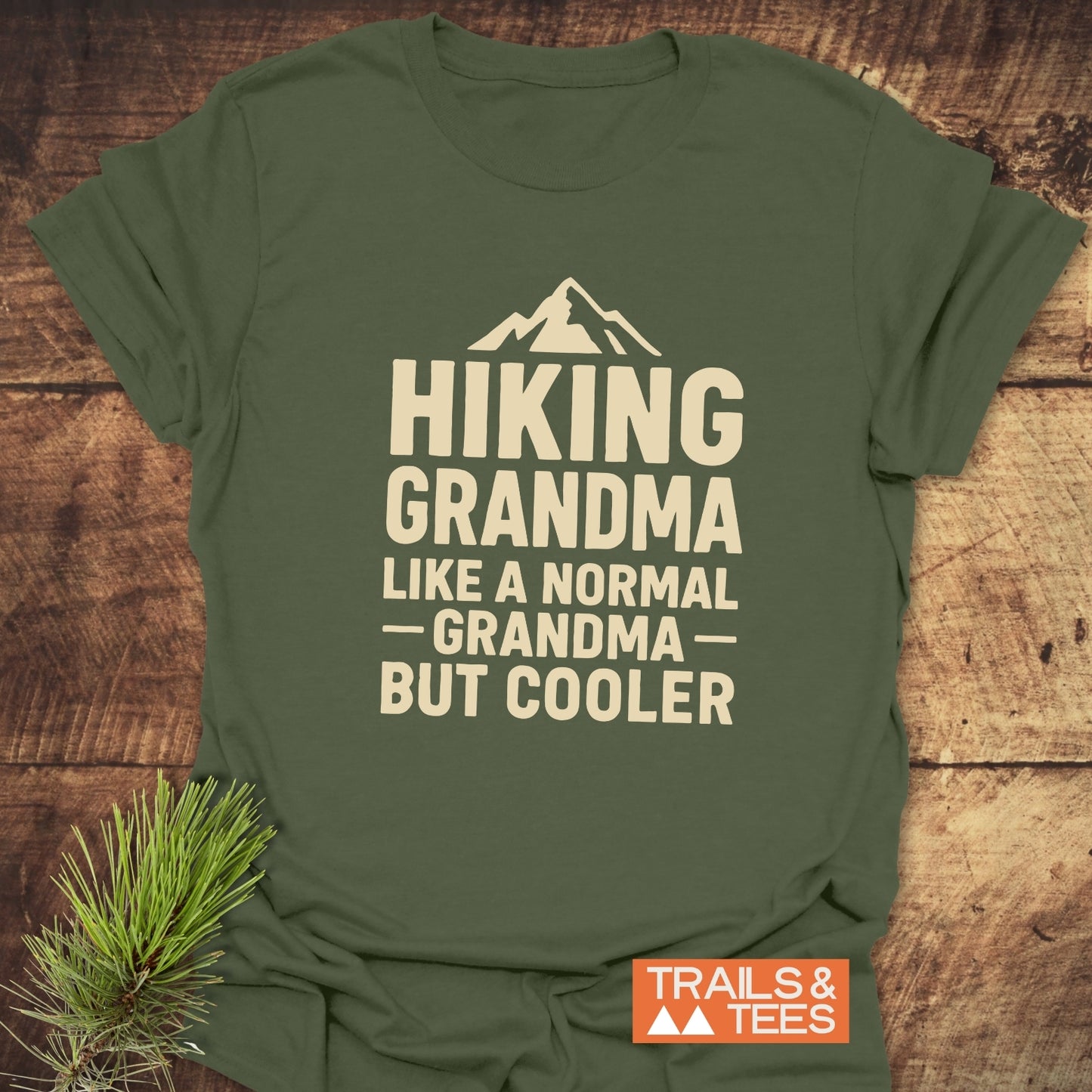 Olive-green Hiking Grandma T-shirt with mountain graphic, crafted from soft cotton for outdoor adventures. Durable print, pre-shrunk, taped shoulders, laid on rustic wood.
