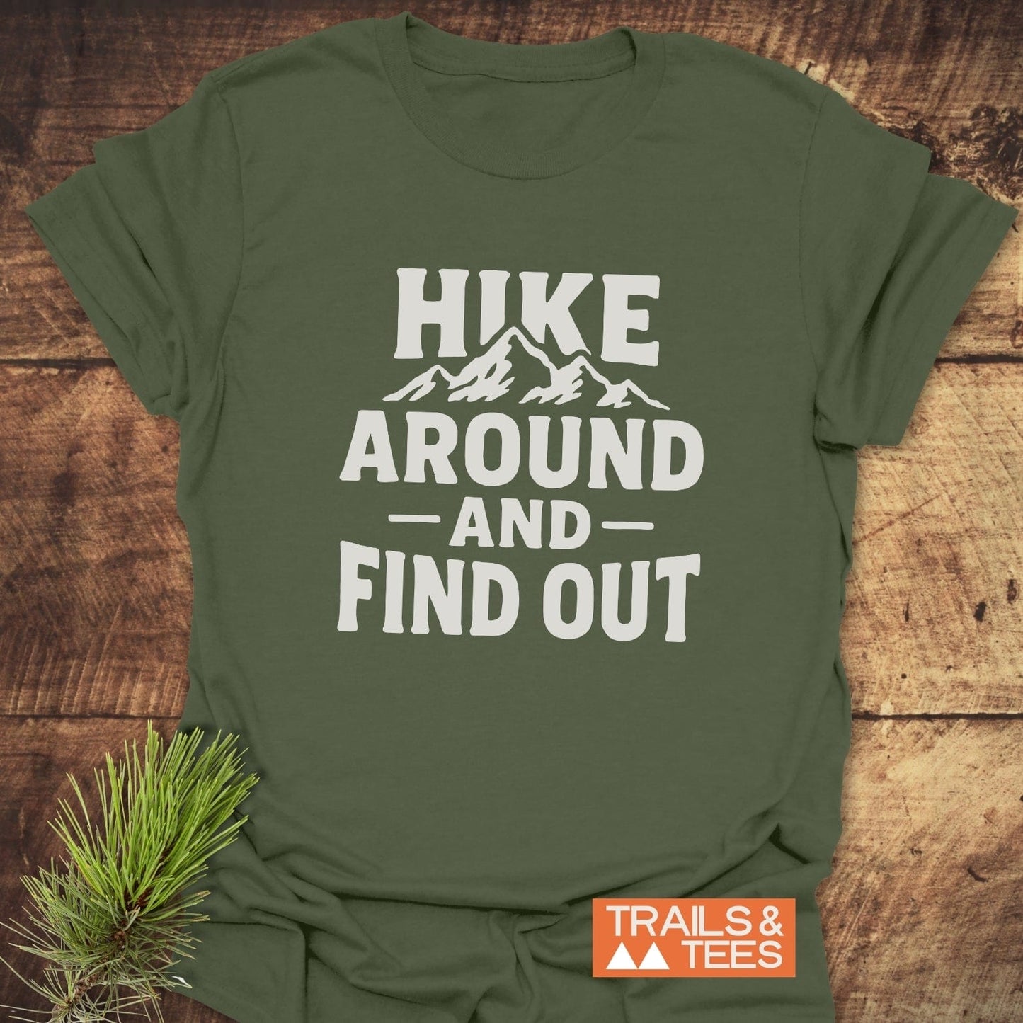 Olive-green T-shirt with mountain silhouette and HIKE AROUND AND FIND OUT text, showcasing outdoor adventure spirit. Includes TRAILS & TEES logo.