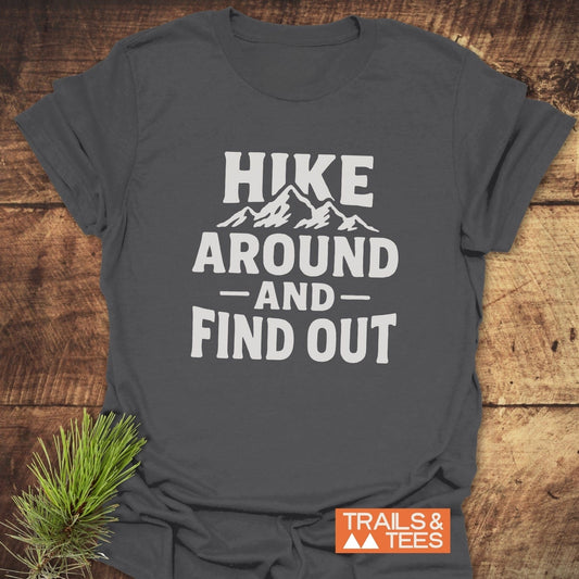 Slate-gray Hike Around And Find Out T-shirt with white mountain print, orange TRAILS & TEES logo, laid on wood with pine bough.