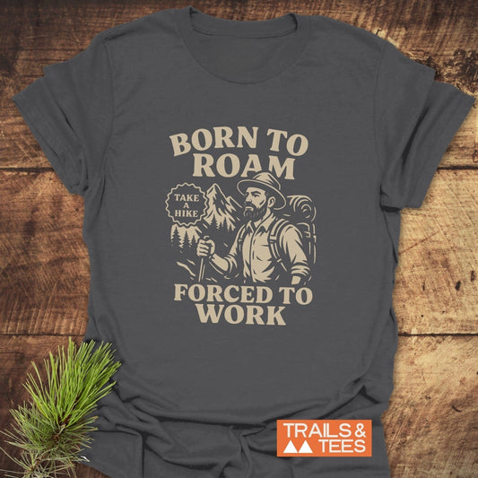 Born To Roam T-Shirt featuring a bearded hiker design with mountains, crafted from soft cotton for comfort and durability, logo in corner, laid on wood surface.