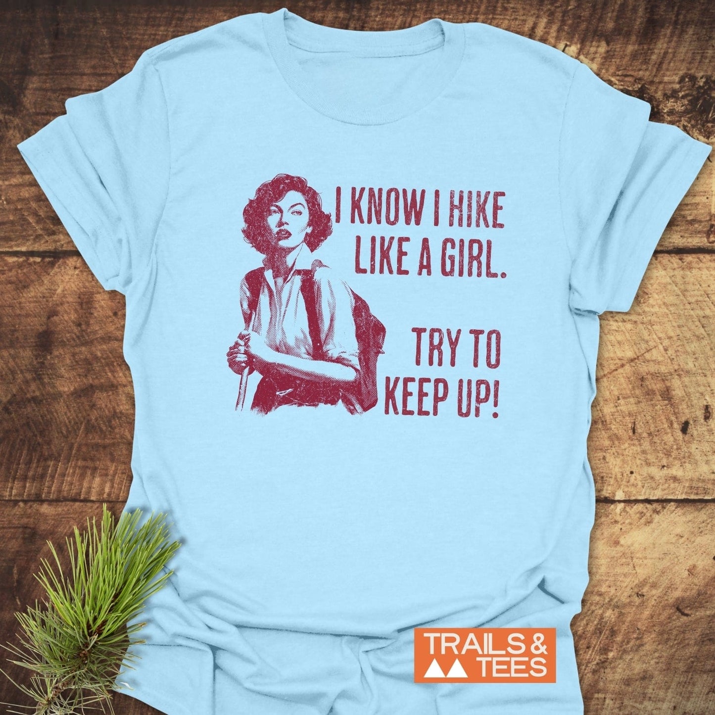 Hike Like A Girl T-Shirt featuring a vintage illustration and slogan, laid on wood with pine sprig; designed for comfort and adventure-focused style.