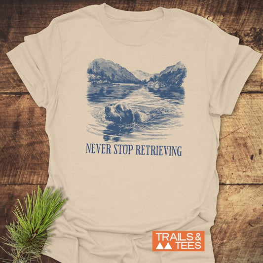 Never Stop... Golden Retriever T-Shirt with blue illustration of a retriever swimming in a lake, slogan below, on a cream-colored cotton fabric, laid on wood.