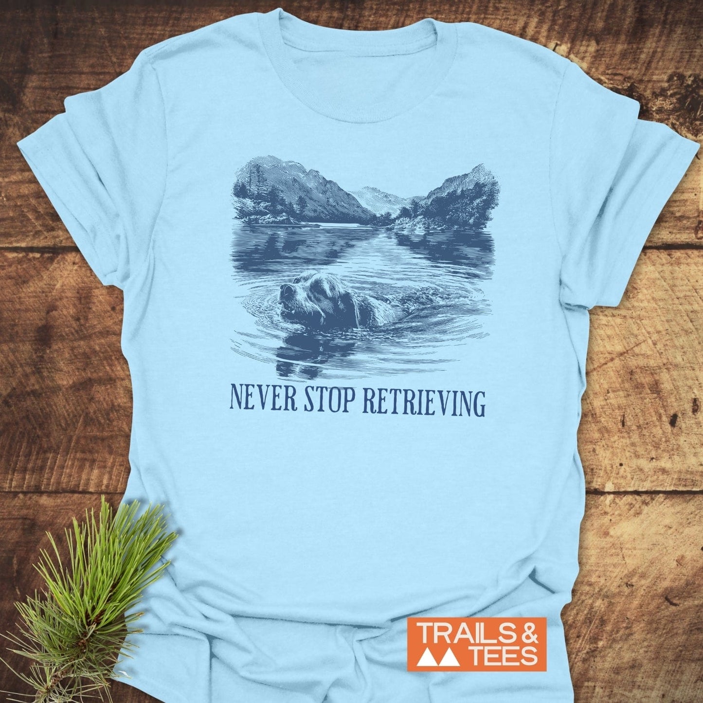 Never Stop... Golden Retriever T-Shirt featuring a retriever swimming design, slogan, and Trails & Tees logo, with pine sprig on wooden surface.