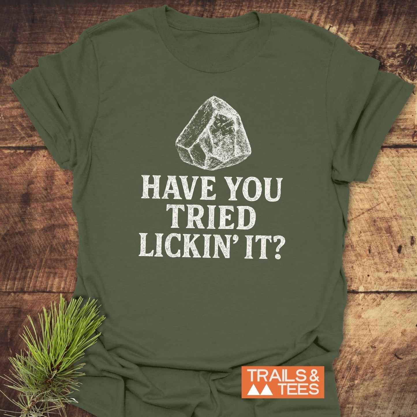 Olive-green 'Have You Tried... Rock Lover T-Shirt' with a rock sketch and slogan, showcasing Trails & Tees logo on a wooden background with pine sprig.