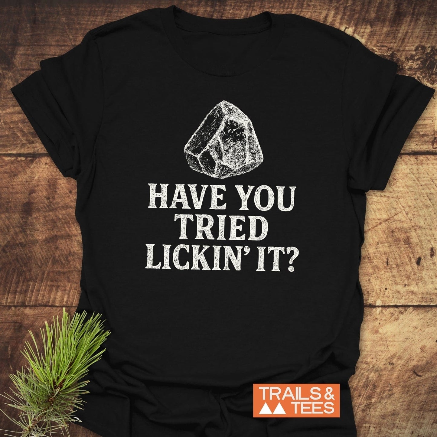 Have You Tried... Rock Lover T-Shirt on wooden surface, featuring a climbing rock sketch and slogan, with Trails & Tees logo, highlighting outdoor adventure spirit.