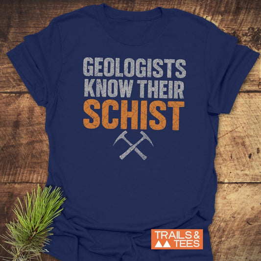 Navy blue Geologists Know Their Schist T-shirt with orange text and crossed rock-hammers, laid on wood. Soft, durable, pre-shrunk cotton for outdoor enthusiasts.