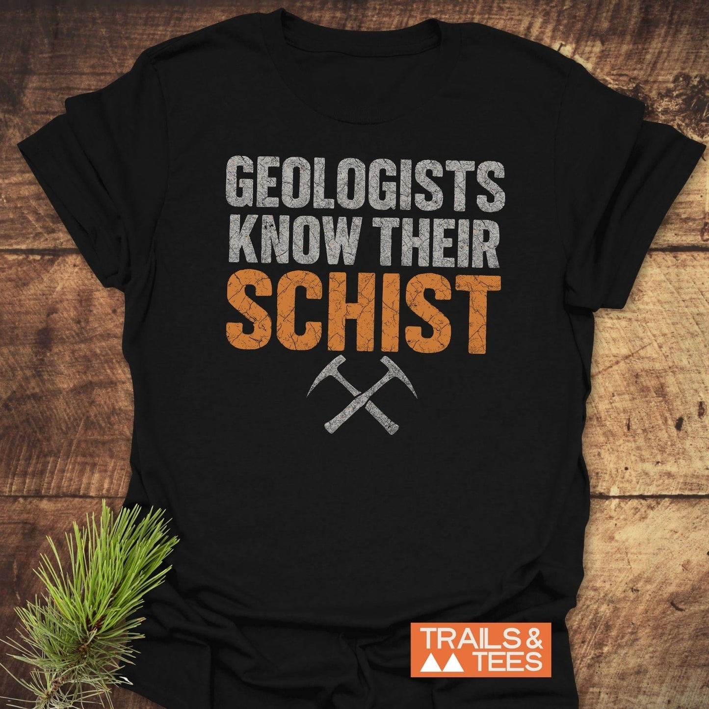 Geologists Know Their Schist T-Shirt with distressed lettering and crossed rock hammers. Black, soft ring-spun cotton, pre-shrunk, durable print, Trails & Tees logo.