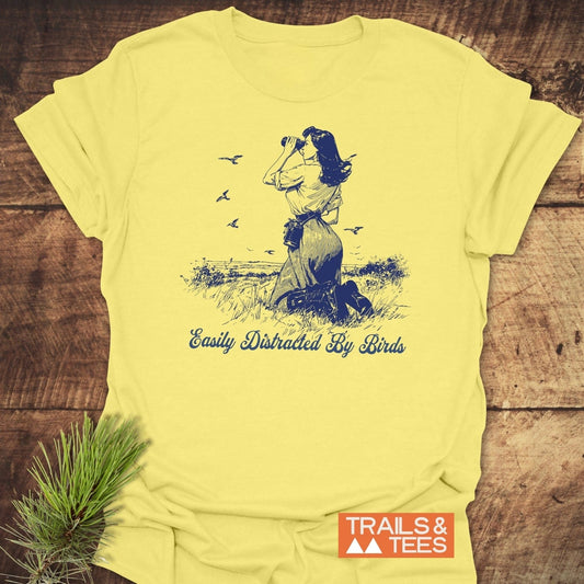 Easily Distracted By Birds T-Shirt featuring a woman with binoculars sketch, slogan, and Trails & Tees logo, laid on a wooden surface with greenery.