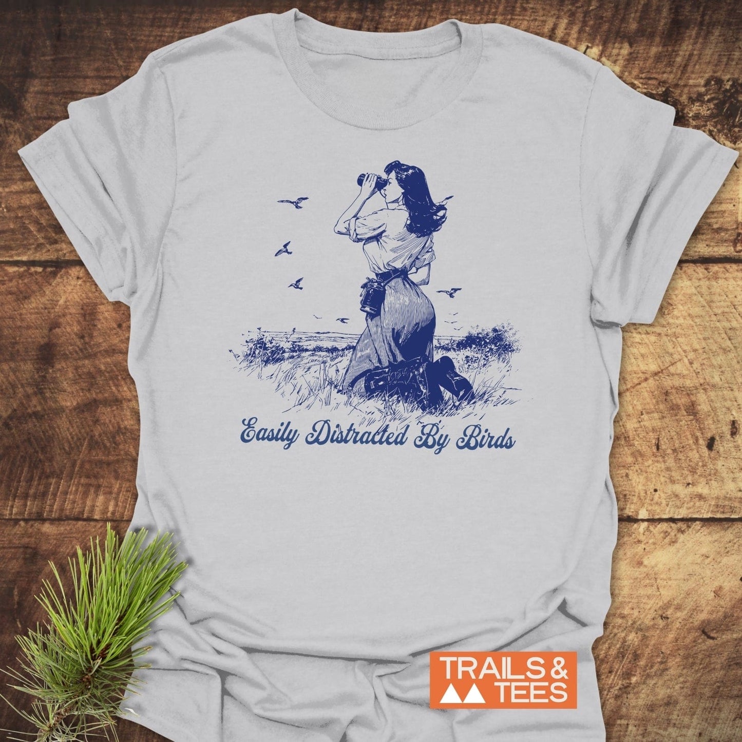 Easily Distracted By Birds T-Shirt featuring a navy illustration of a woman birdwatching, laid on rustic wood. Soft, lightweight cotton, perfect for outdoor enthusiasts.