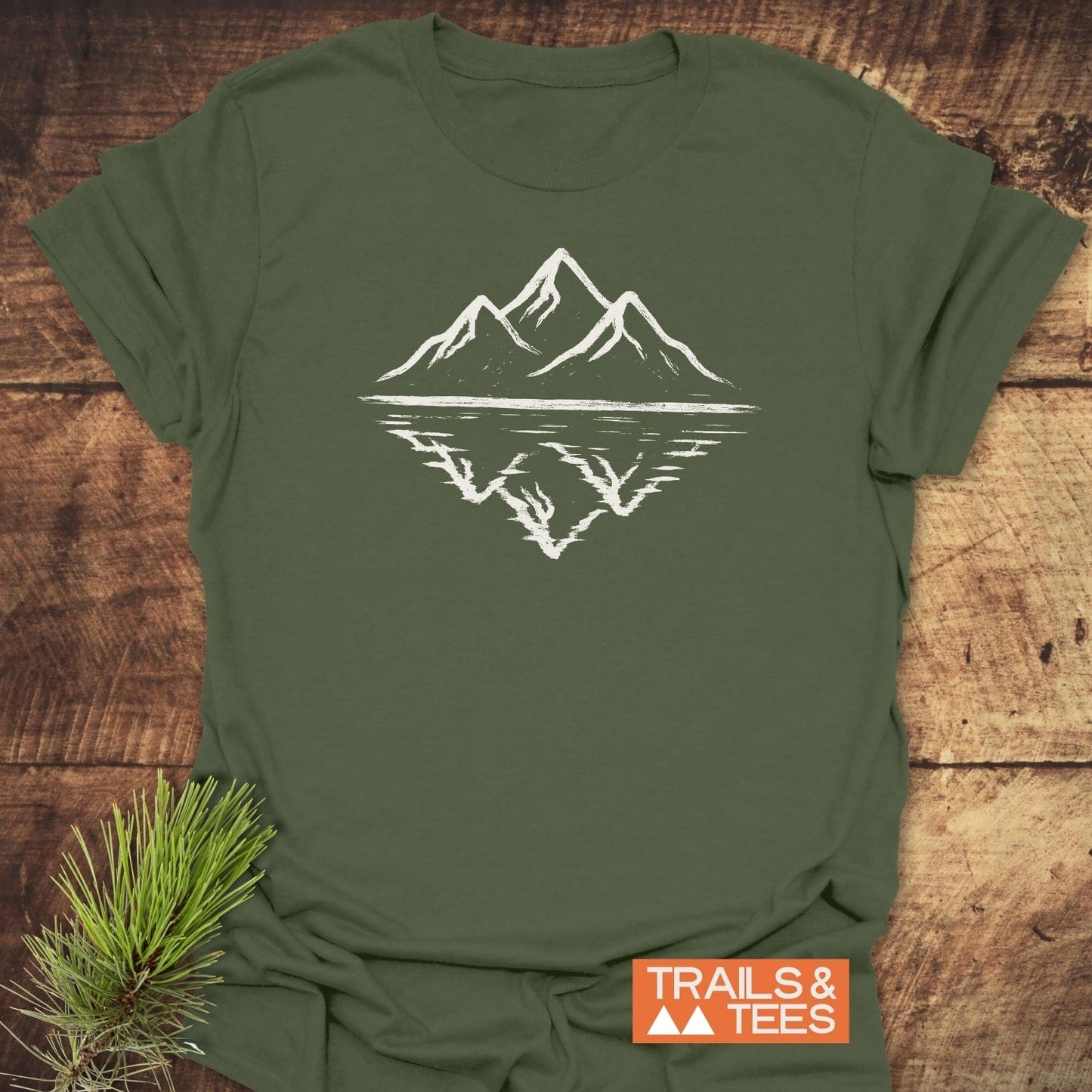 Mountain Reflection T-Shirt laid flat, featuring a white mountain silhouette with reflection. Styled with a pine sprig and an orange TRAILS & TEES label.