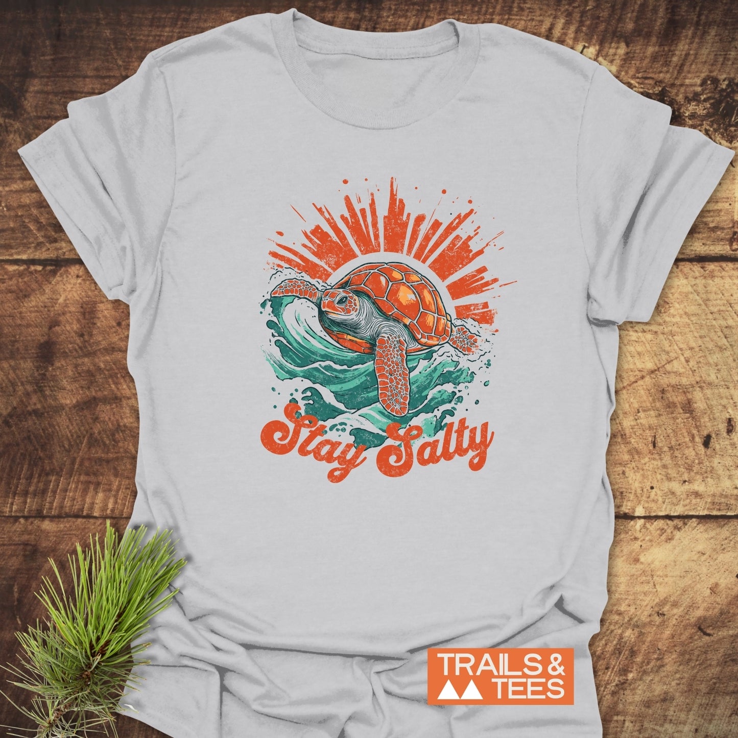 Stay Salty T-Shirt featuring a vintage sea turtle design on a wave, laid on wood with pine sprig, embodying outdoor adventure and comfort by Trails & Tees.