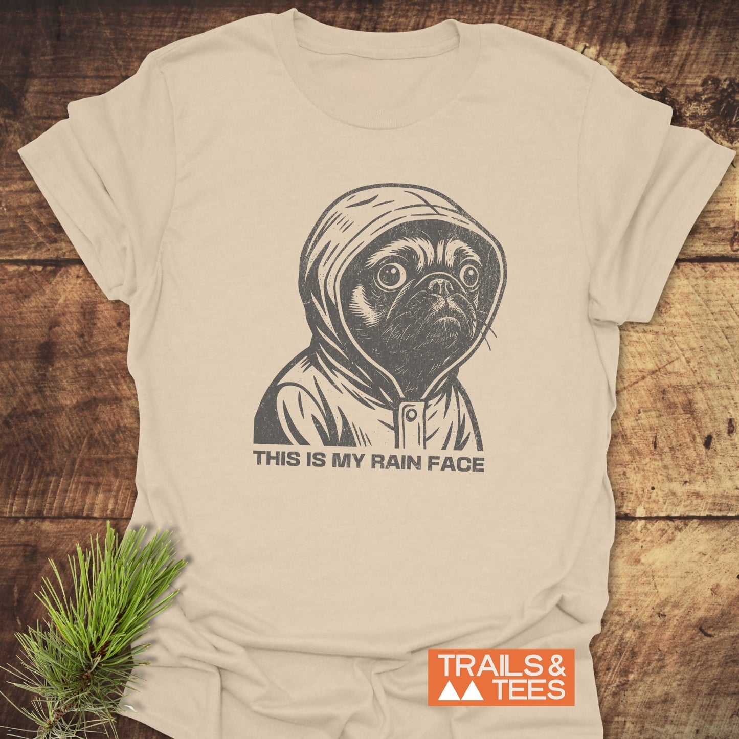 Cream-colored tee featuring a pug in a raincoat sketch with THIS IS MY RAIN FACE, highlighting comfort and durability by Trails & Tees.