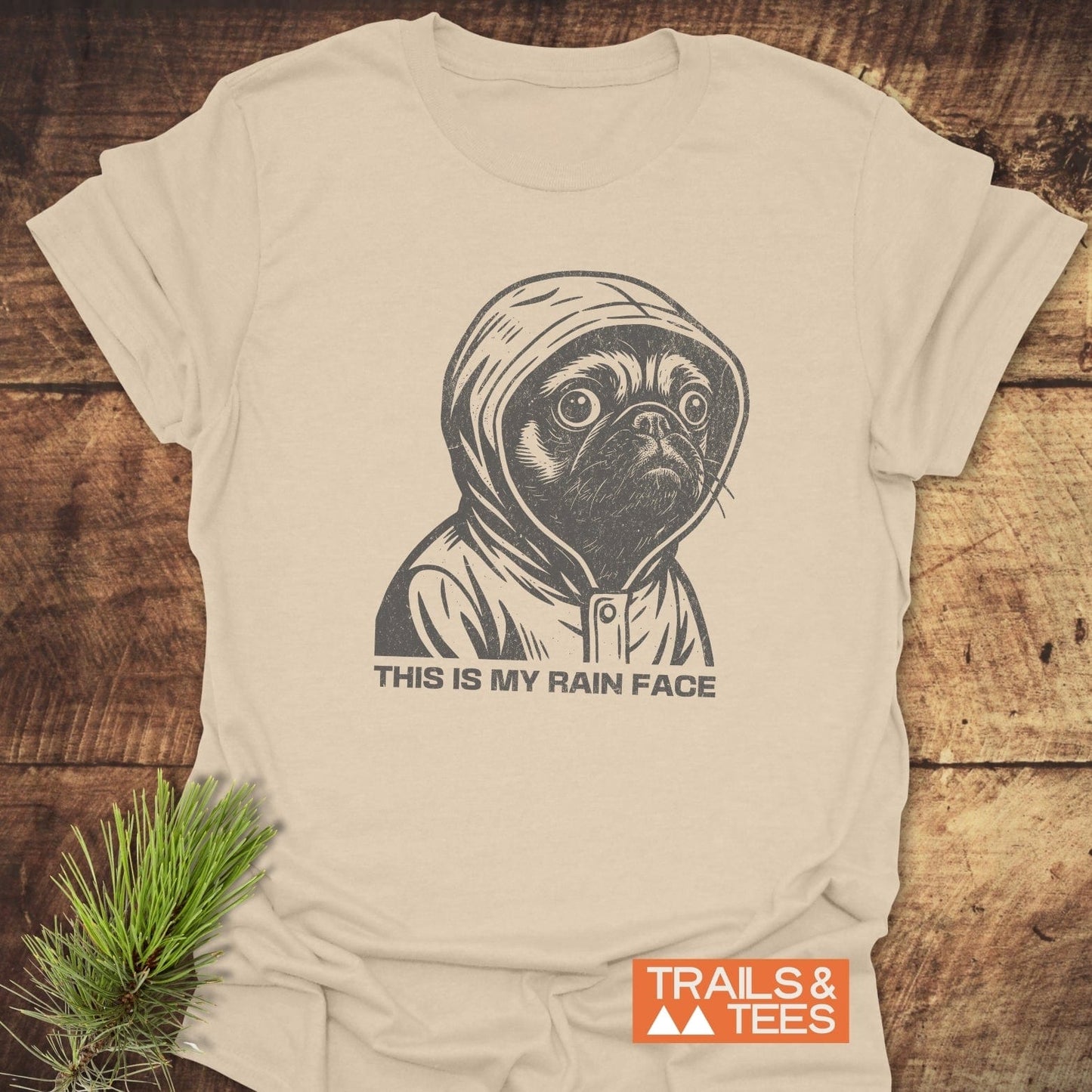 Cream-colored tee featuring a pug in a raincoat sketch with THIS IS MY RAIN FACE, highlighting comfort and durability by Trails & Tees.