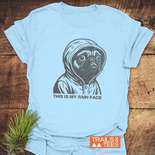 This Is My Rain Face T-Shirt featuring a pug in a raincoat, laid flat with a pine branch accent, embodying comfort and outdoor adventure spirit.