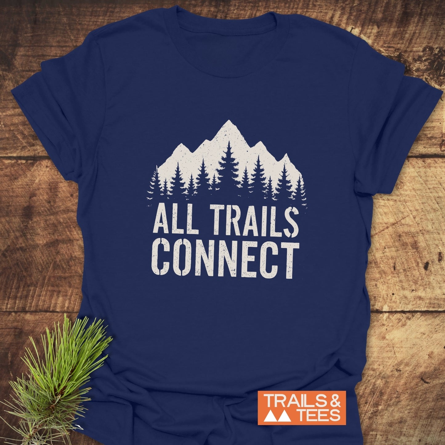 All Trails Connect T-Shirt featuring a white mountain graphic with pine trees and slogan on navy-blue. Includes orange TRAILS & TEES label and pine sprig.