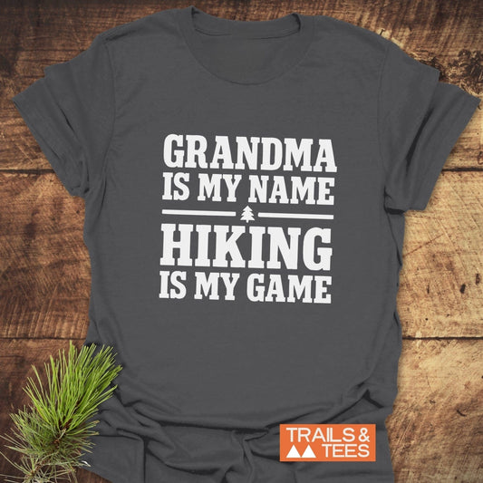 Grandma Hiking T-Shirt on wood surface, featuring bold white lettering and a pine tree icon, with TRAILS & TEES logo and pine sprig beside it.