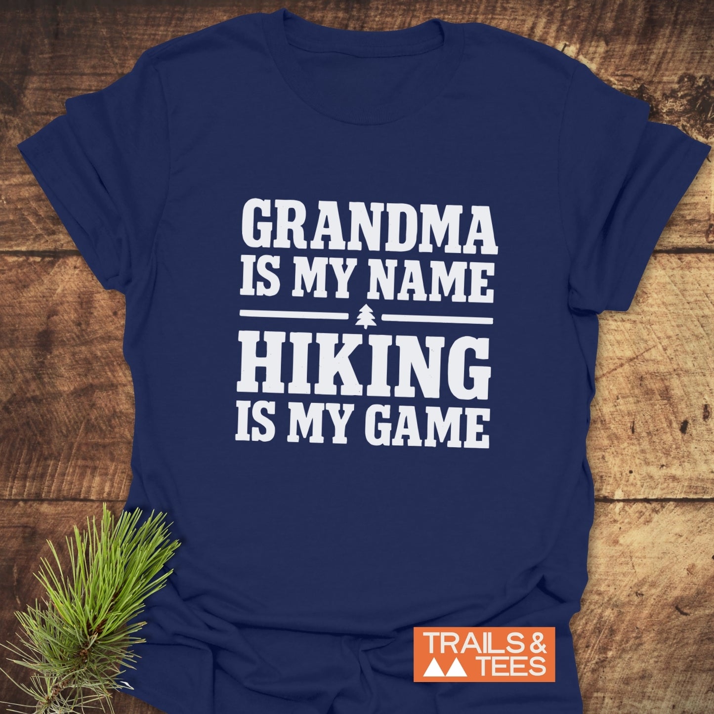 Navy-blue Grandma Hiking T-Shirt on wooden surface, featuring bold white text and pine-tree graphic, styled with a pine sprig and Trails & Tees logo.