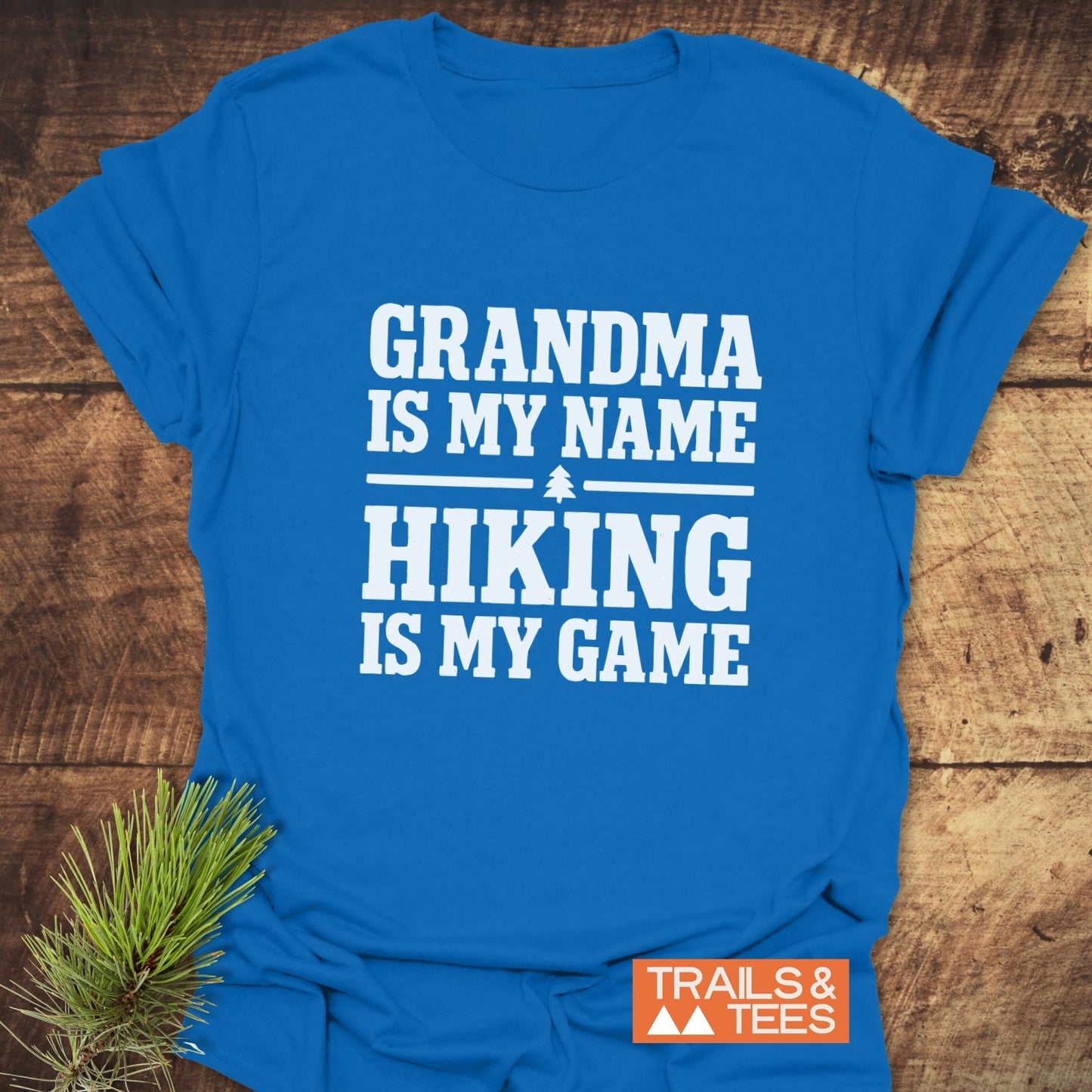 Grandma Hiking T-Shirt with GRANDMA IS MY NAME, HIKING IS MY GAME slogan, tree icon, and TRAILS & TEES logo, displayed on rustic wood with pine branch.