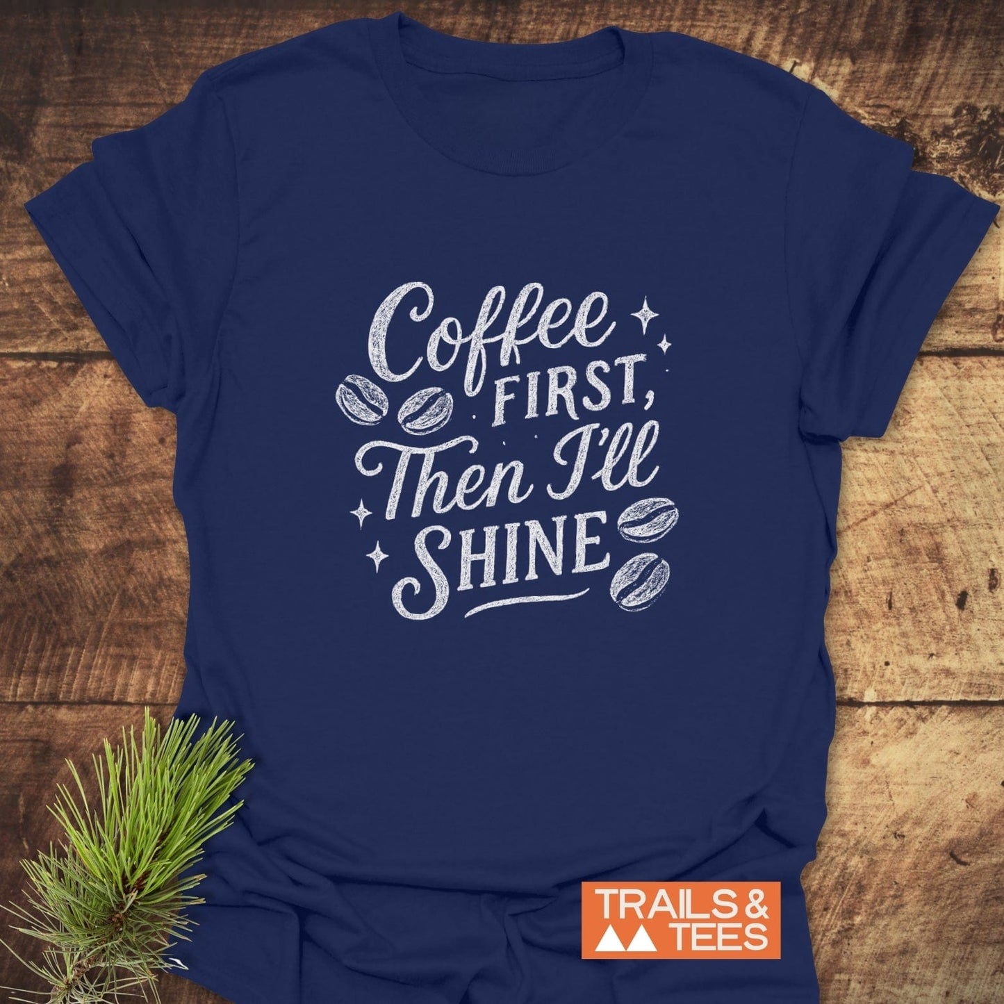 Navy-blue Coffee First T-Shirt with white chalk-style lettering, coffee-bean illustrations, and sparkle accents, featuring TRAILS & TEES logo on wooden surface.
