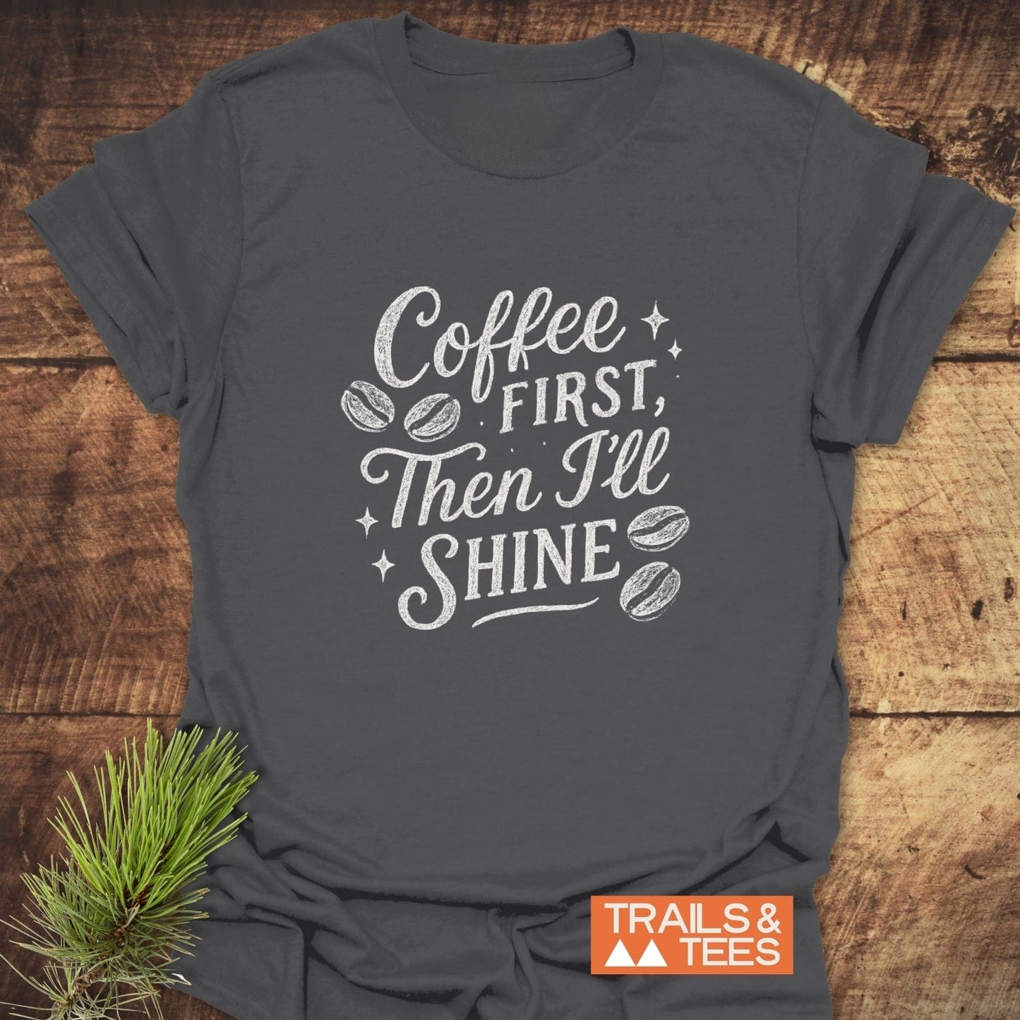 Charcoal-gray Coffee First T-Shirt with white text and coffee bean icons, featuring a small TRAILS & TEES logo, styled with a pine sprig.
