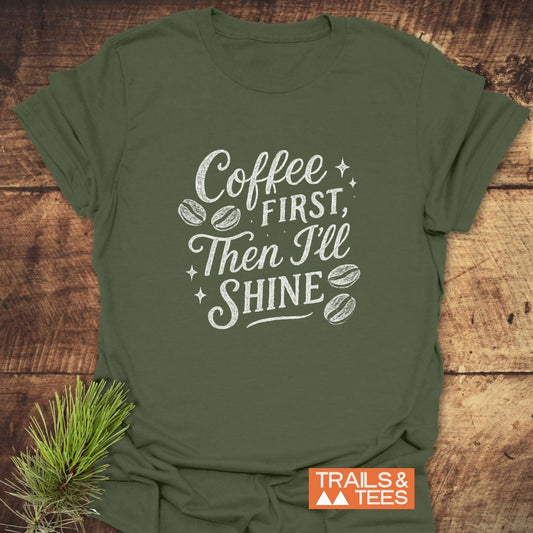 Forest-green Coffee First T-Shirt with white slogan and starbursts, styled on wood with pine sprig; Trails & Tees logo in corner.
